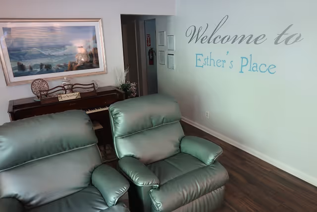 Interior view of a senior living facility named Esther's Place showing two green recliner chairs in the foreground, a piano with decorative items on top against the wall, a framed painting of a seascape above the piano, and a wall with the text 'Welcome to Esther's Place'.