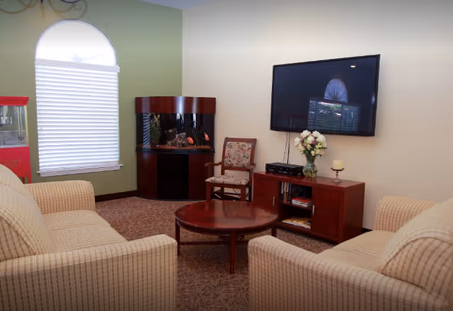 A cozy living room with two beige striped armchairs facing a round wooden coffee table. A wooden chair with floral upholstery is placed near a corner fish tank. A flat-screen TV is mounted on the wall above a wooden cabinet holding books, a vase with flowers, and a candle. A window with a white blind and an arched top is on the left wall.