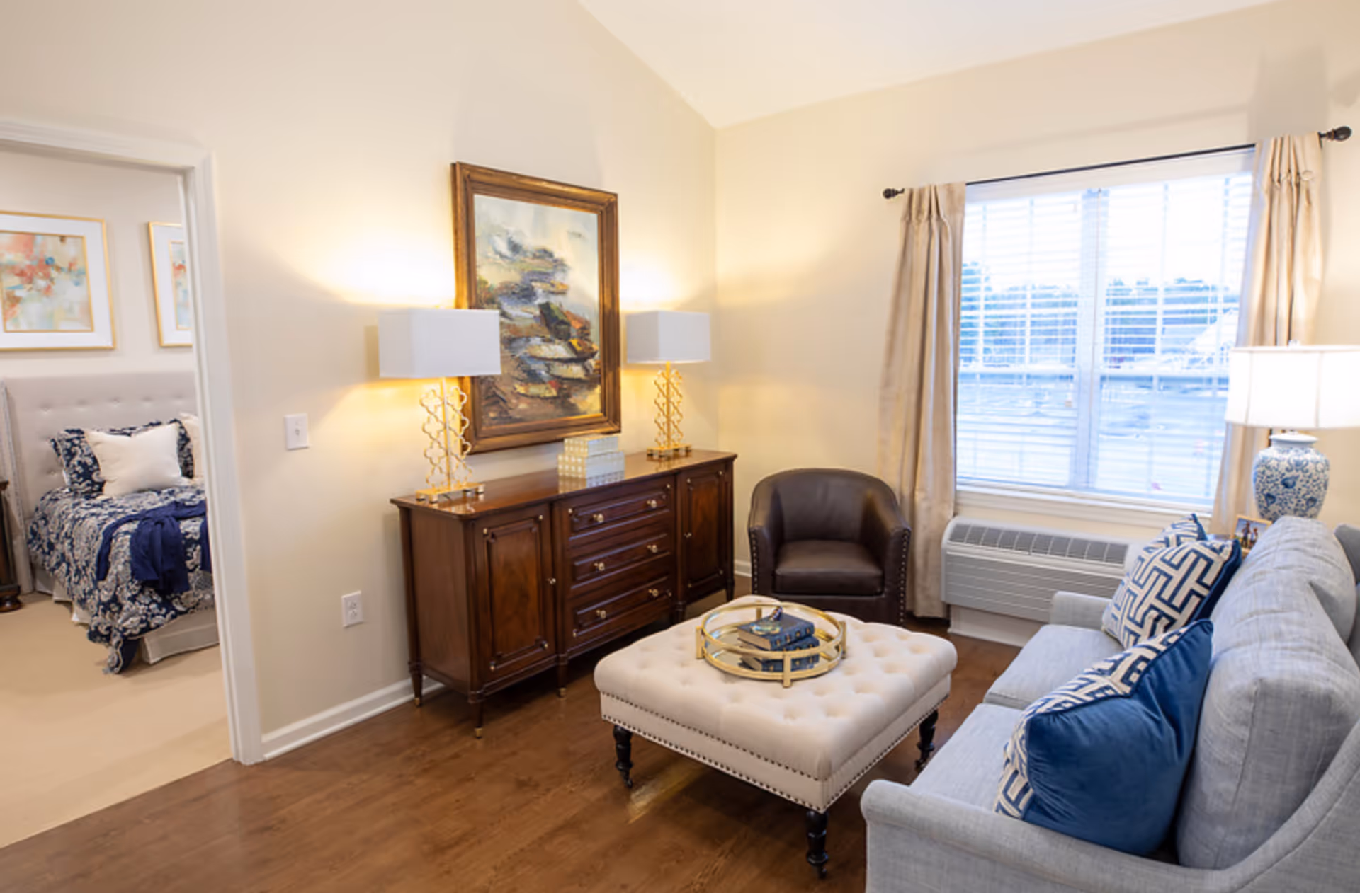 A cozy living room with a light gray sofa adorned with blue and patterned pillows, a tufted ottoman with a decorative tray, a dark brown leather armchair, and a wooden sideboard with two lamps and a framed painting above it. A window with beige curtains lets in natural light. Through an open doorway, a bedroom with a bed dressed in blue and white bedding is visible.