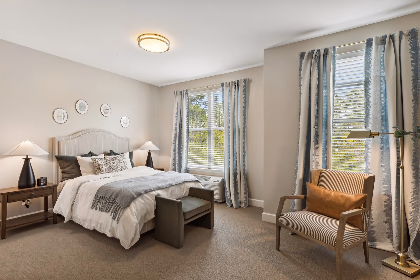 A cozy bedroom with a large bed featuring white and gray bedding, two bedside tables with lamps, a small bench at the foot of the bed, two windows with blue and white curtains, and a striped armchair with a brown cushion next to a floor lamp.