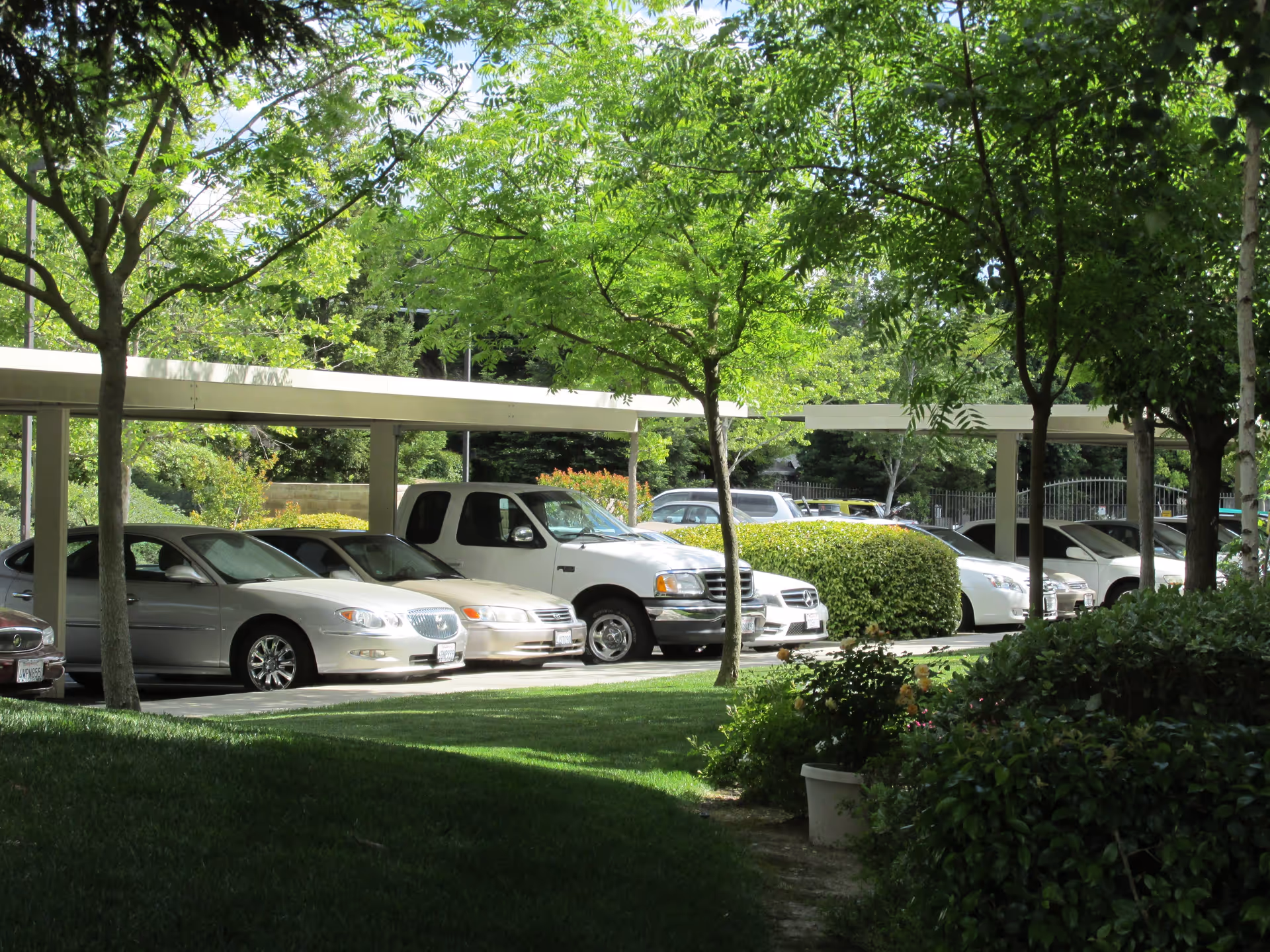 A shaded parking area with several cars and a white pickup truck parked under a beige carport. The parking lot is surrounded by green trees, bushes, and well-maintained grass, creating a peaceful outdoor environment.