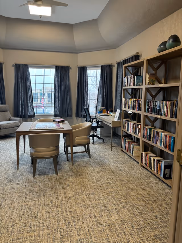 A cozy room with a table and four chairs in the center, a bookshelf filled with books on the right side, a desk with a chair near the windows, and a comfortable armchair in the corner. The room has large windows with dark curtains and a ceiling fan with a light fixture.