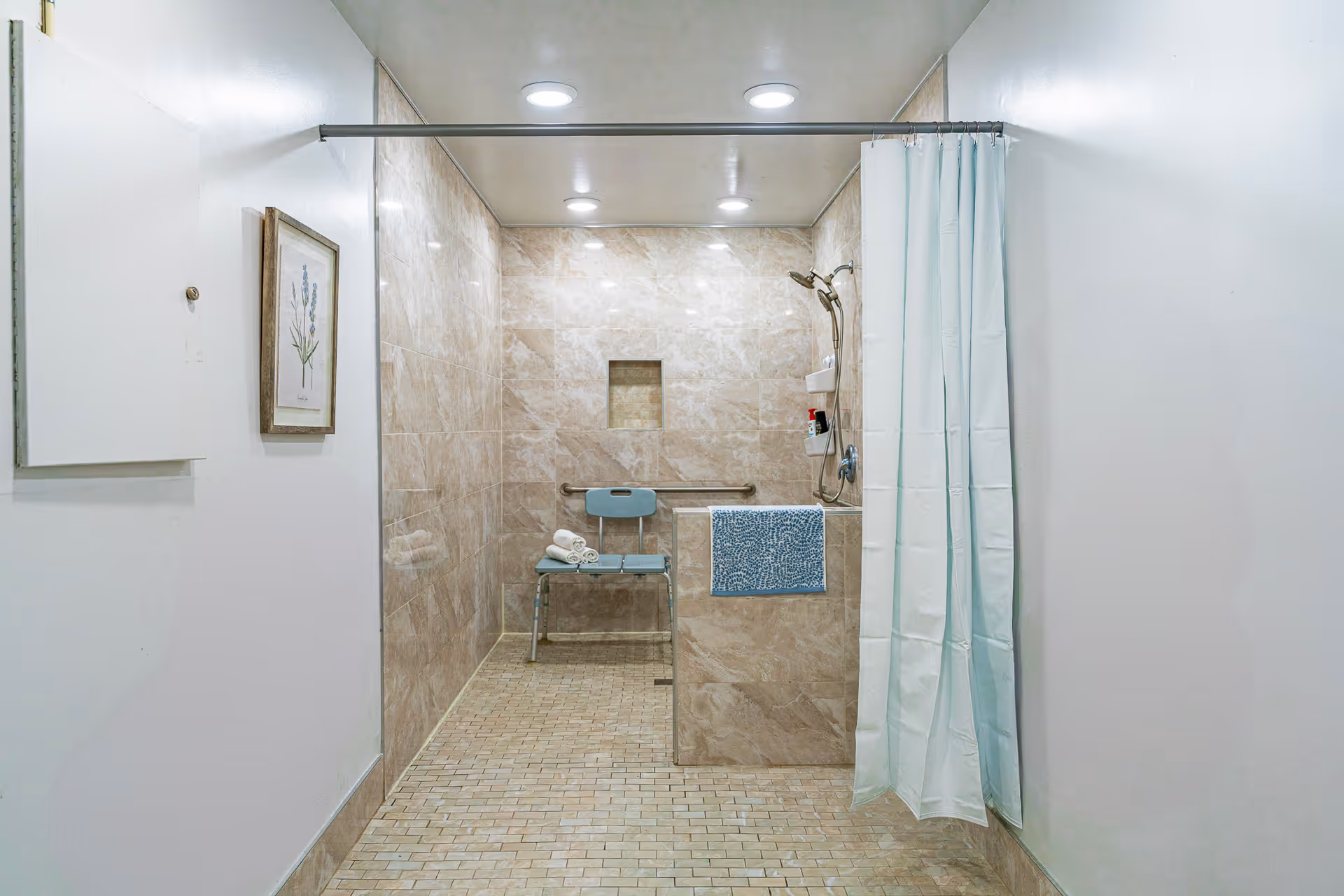 A spacious, well-lit shower area with beige tiled walls and floor. The shower features a blue shower chair with rolled white towels on it, a handheld showerhead, a grab bar, and a small built-in shelf with toiletries. A white shower curtain is partially drawn to the right side. There is a framed botanical print on the left wall outside the shower area.