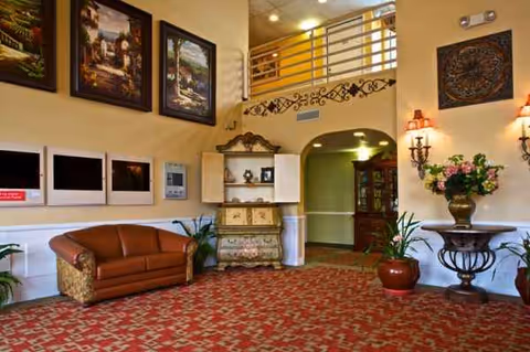 A warmly furnished interior lobby with a red patterned carpet, sofa, artwork, plants, and decorative furniture.