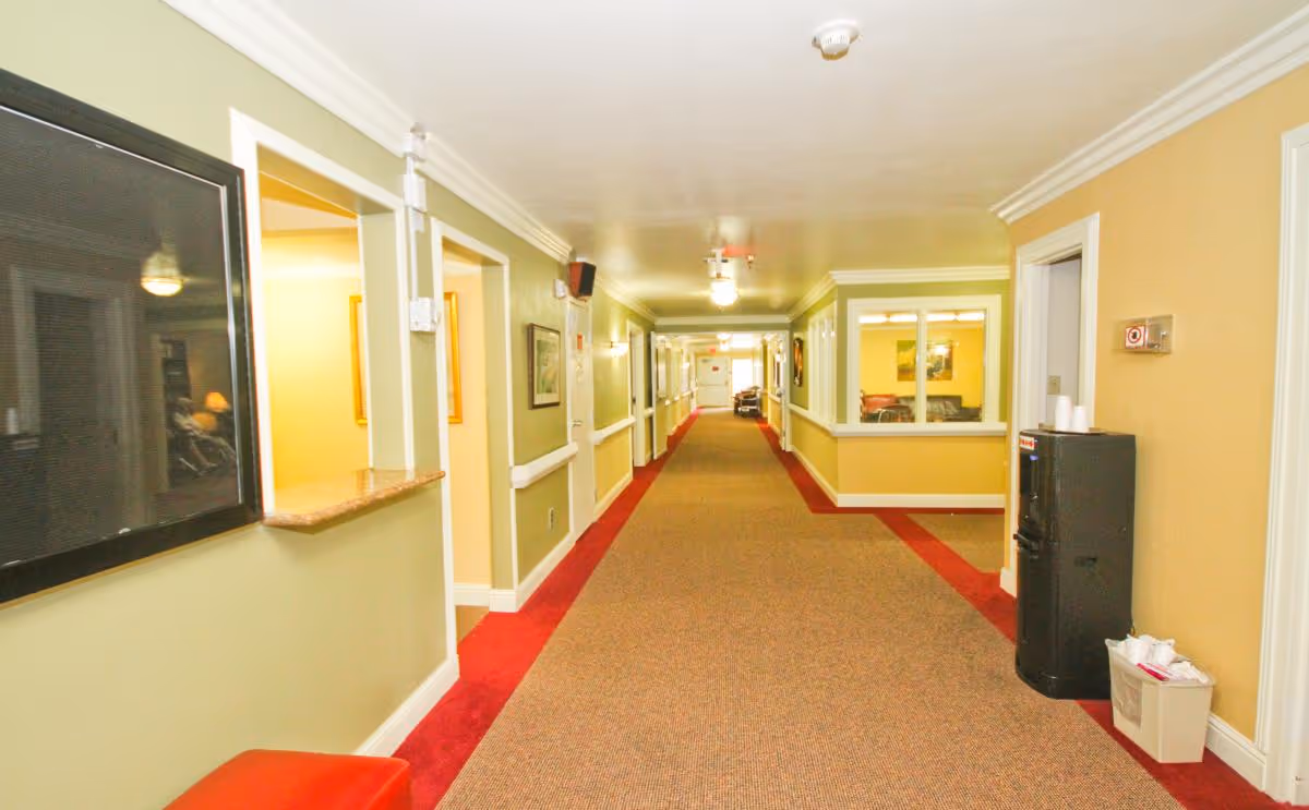 Carpeted, well-lit interior hallway of an assisted living facility with seating areas, windows into rooms, and a water cooler.