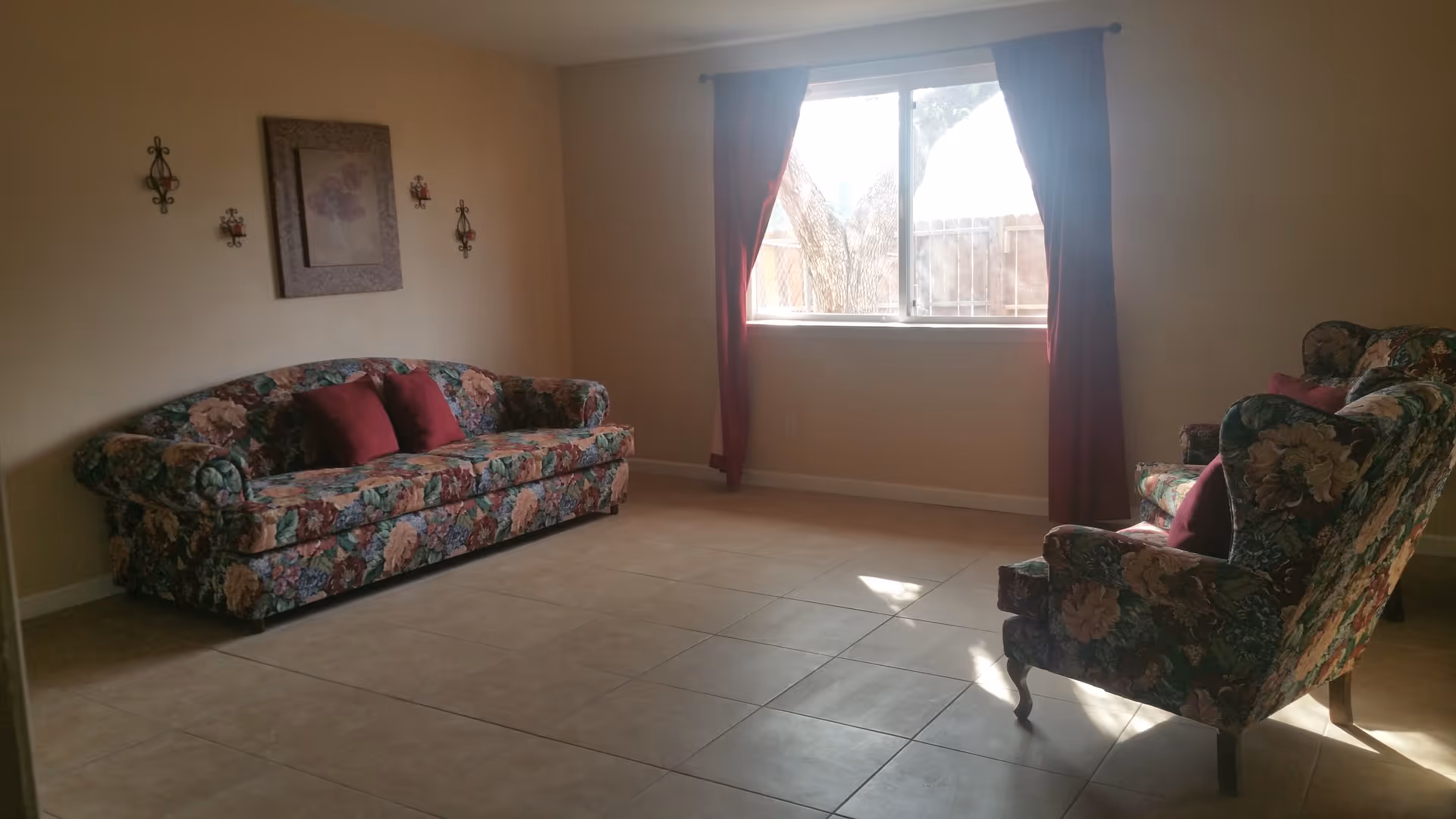 A living room with a floral patterned sofa and matching armchair, both with red accent pillows. The room has beige tiled flooring, beige walls, a window with red curtains letting in sunlight, and decorative wall hangings including a framed picture and candle holders.