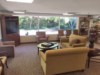 A cozy living room area with large windows showing a green outdoor space. The room contains beige and brown upholstered armchairs and sofas arranged around a round coffee table with a decorative tray. There is a wooden cabinet with a TV on the left side and a small side table with a plant on the right. The carpet is patterned, and the room has a warm, inviting atmosphere.