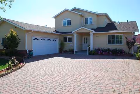 Two-story beige house with a brick-pattern driveway, front entry, and blooming flower beds.