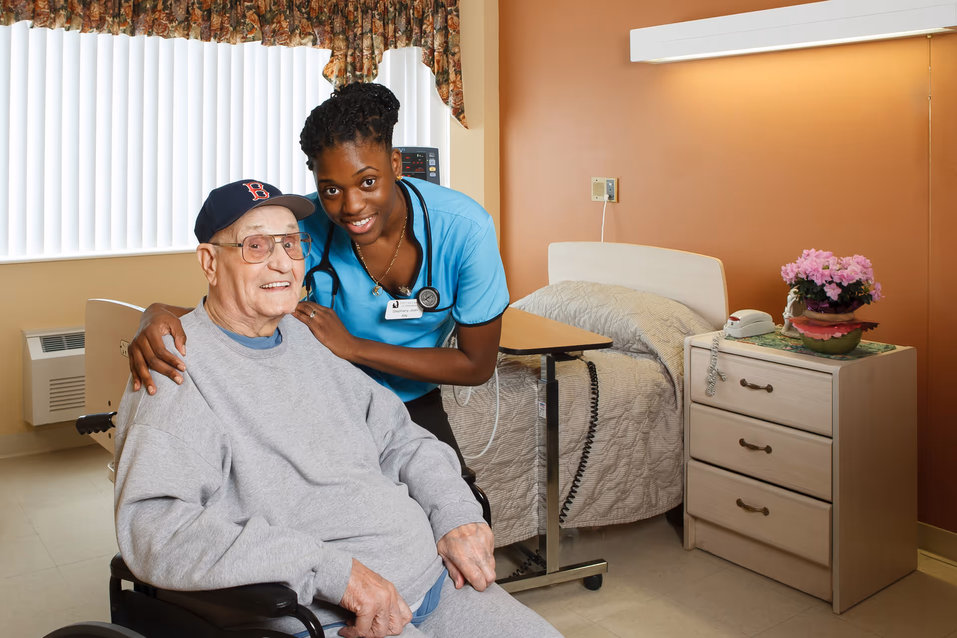 A smiling elderly man in a wheelchair wearing a gray sweatshirt and a Boston Red Sox cap is accompanied by a smiling female healthcare worker in blue scrubs with a stethoscope around her neck. They are in a well-lit room with a bed, bedside table with a phone and flowers, and a window with vertical blinds and floral valance.