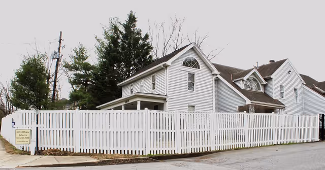Exterior view of a two-story white house with multiple windows and a white picket fence surrounding the property. There are some trees behind the house and a sign on the fence that reads AlfredHouse Elder Care, Inc.