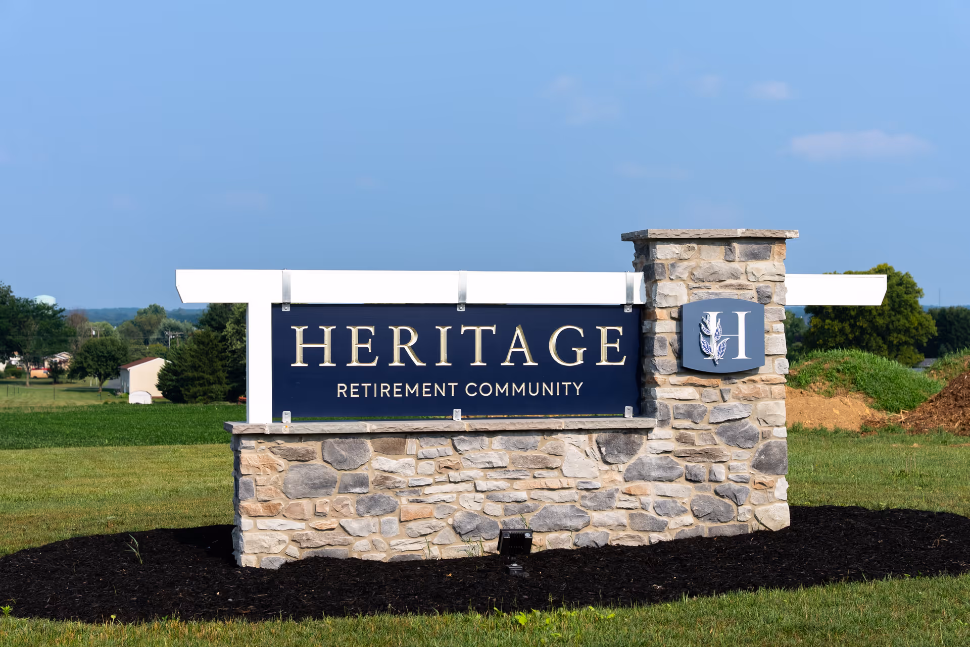 Stone and wood sign for Heritage Retirement Community on a grassy area with trees and a clear blue sky in the background.