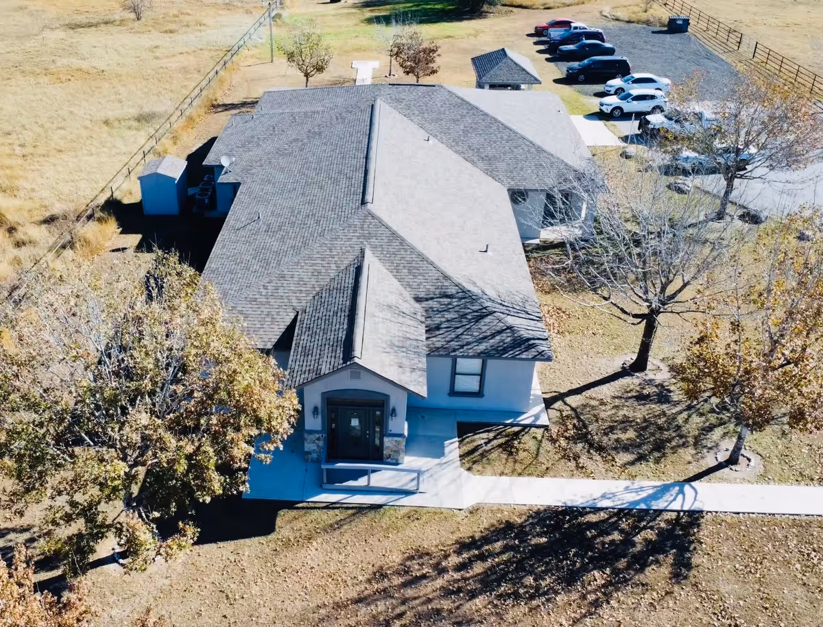 Aerial view of a single-story building with a gray shingled roof surrounded by trees with autumn foliage. There is a concrete walkway leading to the front entrance, and a parking area with several cars is visible in the background. The building is situated in a grassy area with a fence running along one side.