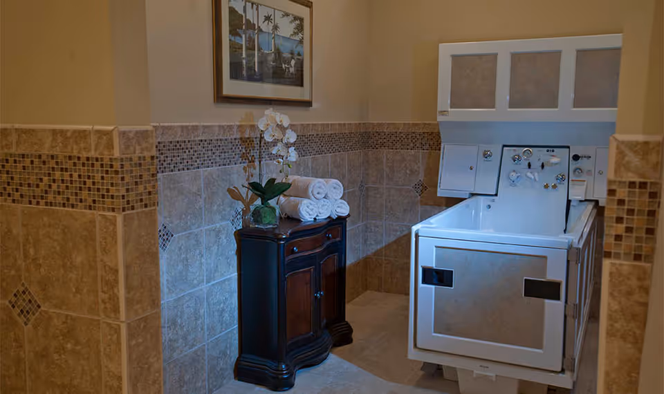 Tiled bathing room with a walk-in tub on the right, a small wooden cabinet topped with rolled towels and an orchid, and framed artwork on the wall.