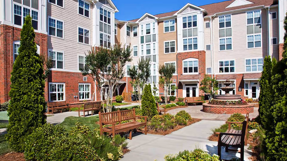 Outdoor courtyard area of a senior living facility with multiple wooden benches, landscaped greenery, small trees, and a central brick fountain. The courtyard is surrounded by a multi-story building with large windows and a mix of brick and siding exterior.