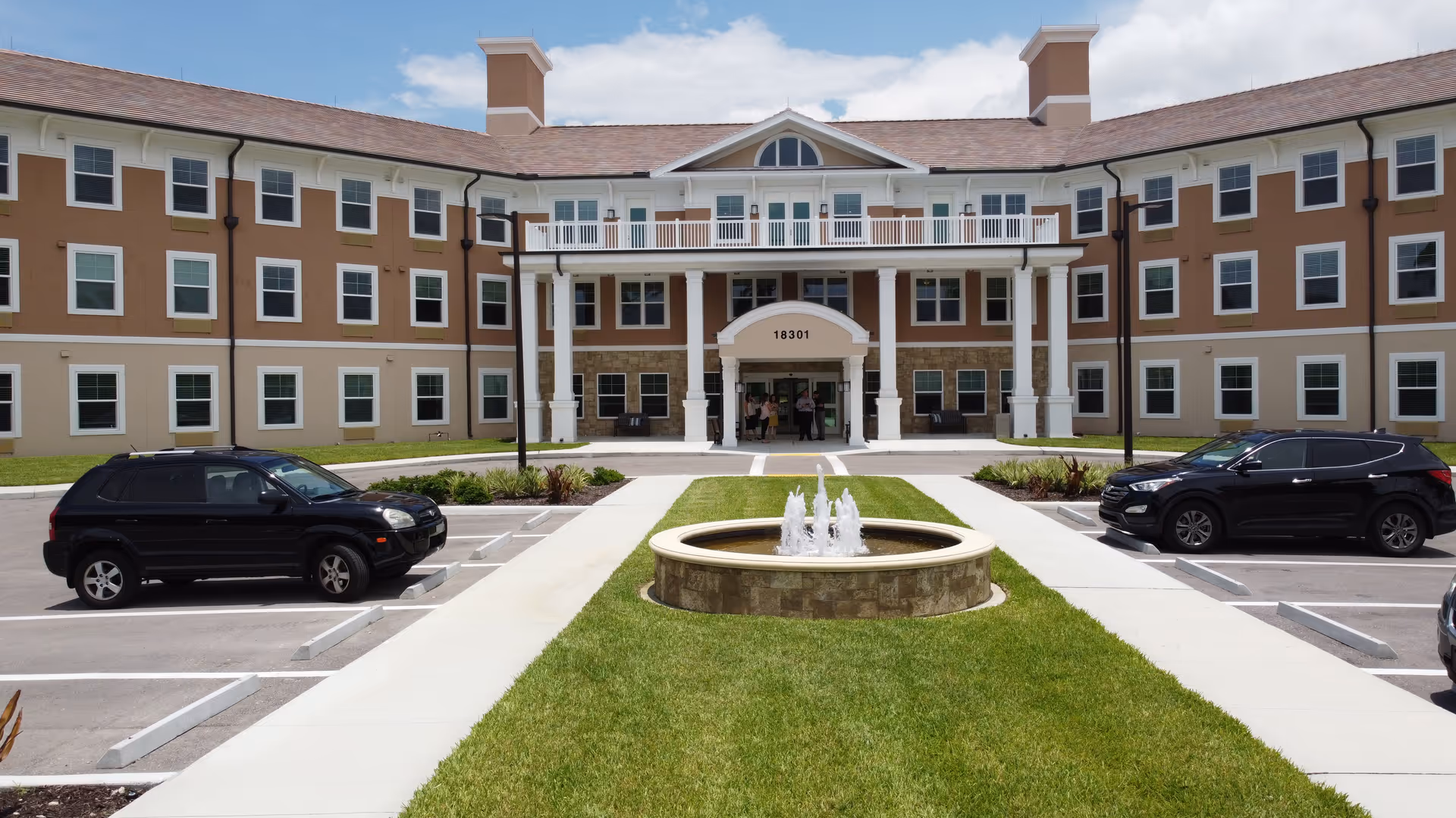 Front exterior view of a senior living facility building with a central entrance featuring white columns and a covered porch. There is a circular water fountain in the foreground surrounded by green grass, with parking spaces and two black SUVs on either side of the walkway leading to the entrance. The building has three stories with multiple windows and a beige and brown color scheme.