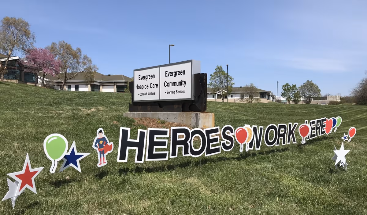 Outdoor grassy area with a sign for Evergreen Hospice Care and Evergreen Community. In front of the sign, large letters and decorations on the grass spell out 'HEROES WORK HERE' with stars, balloons, and a superhero figure.