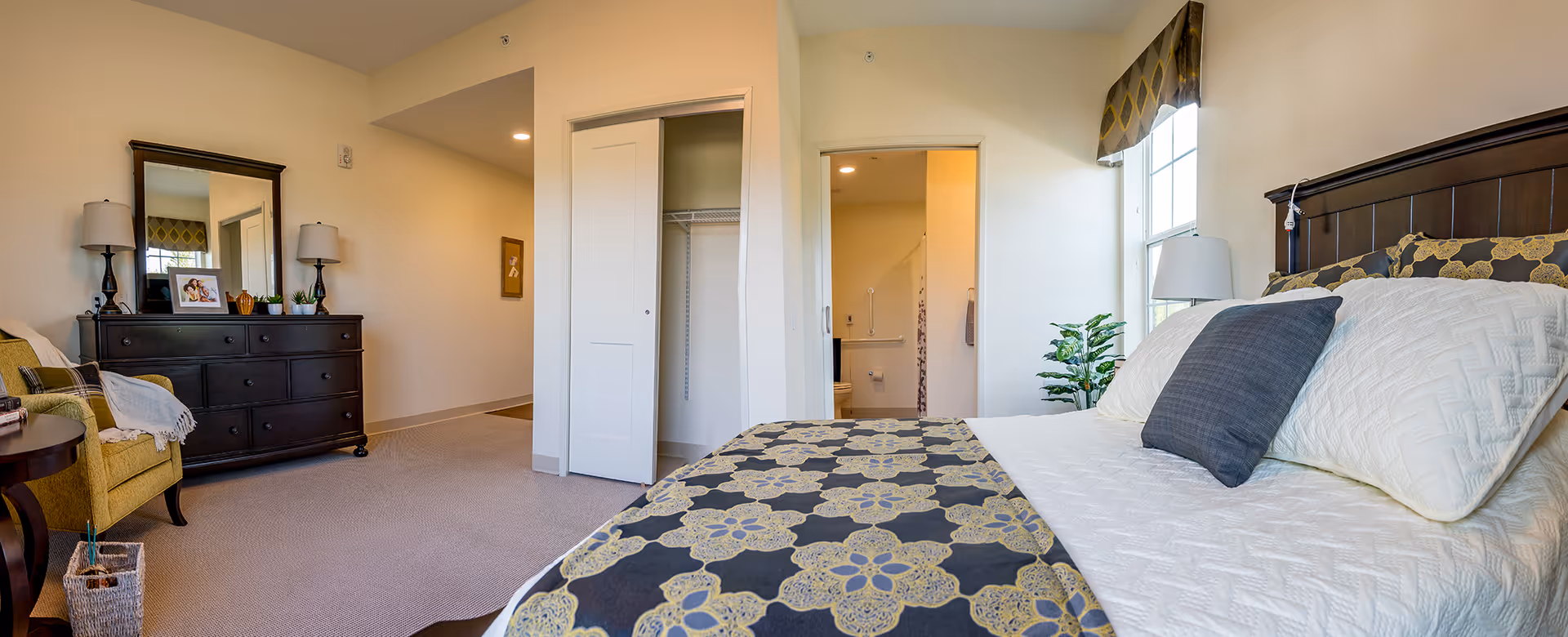 A bright and spacious bedroom in Walnut Crossing featuring a large bed with white and patterned bedding, a dark wooden headboard, and a window with a patterned valance. To the left, there is a green armchair with a plaid pillow and a dark wooden dresser with a large mirror and two lamps. An open closet with white sliding doors and an open doorway leading to a bathroom are visible in the background. The room has beige walls and carpeted flooring.