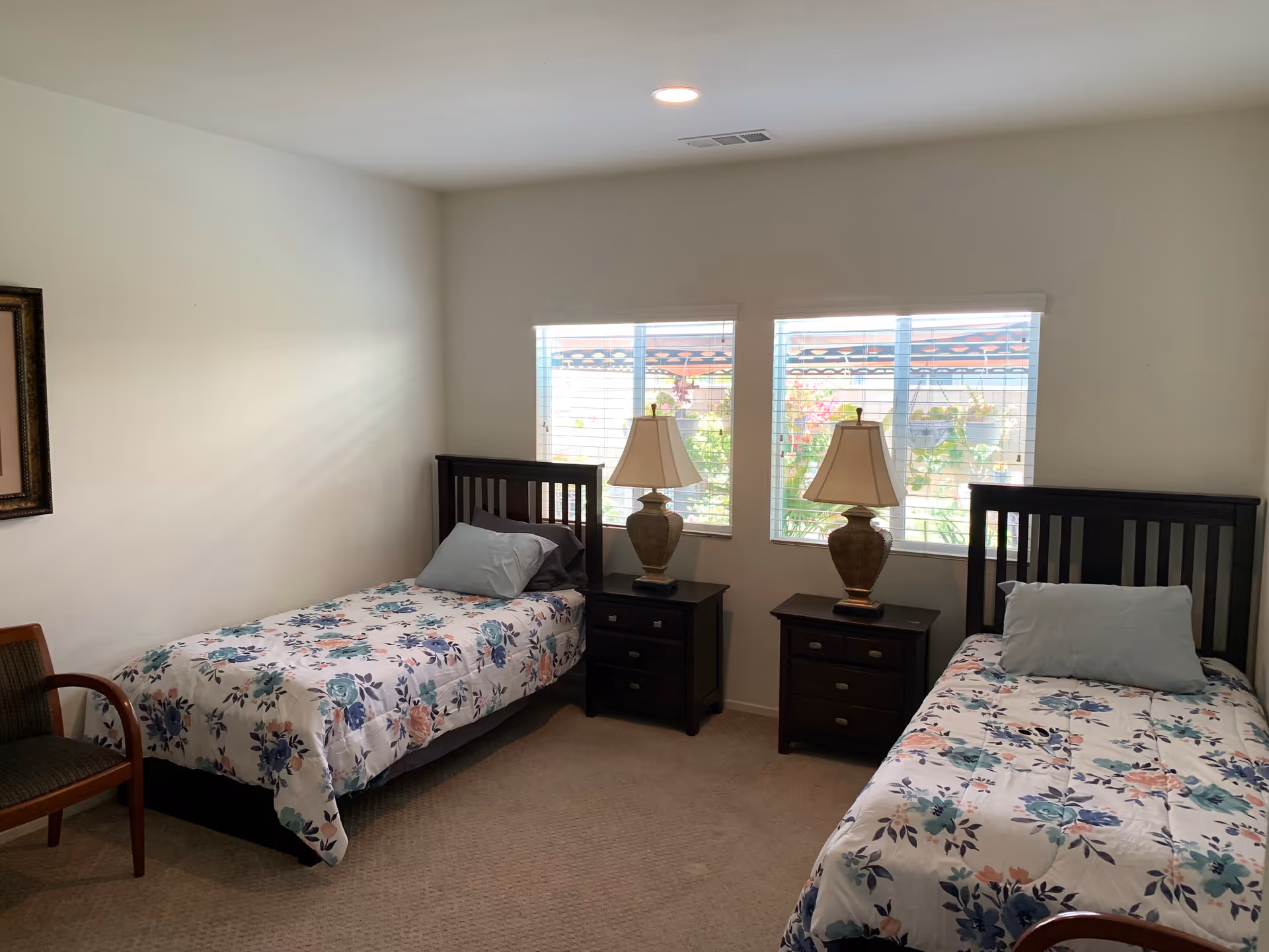 A bright twin bedroom with two single beds, floral bedspreads, two nightstands with lamps, and windows letting in natural light.