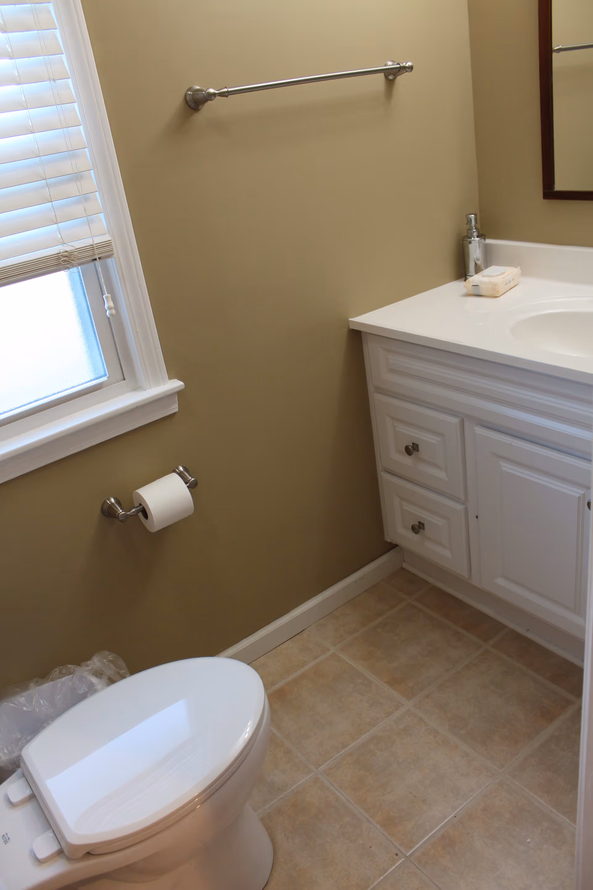Small bathroom with a toilet, vanity and sink, towel bar, toilet paper holder, and a window with blinds.