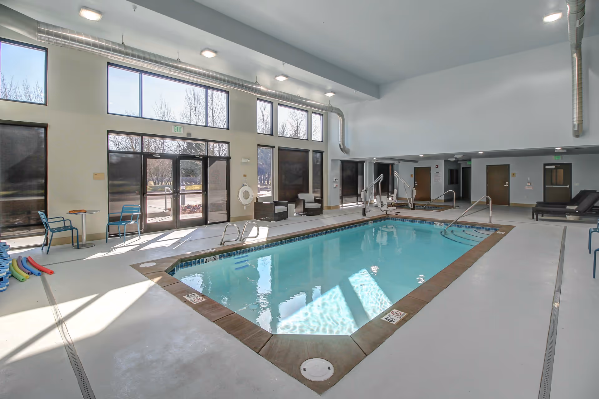 Indoor community swimming pool in a bright room with large windows, seating, and an accessible pool lift.