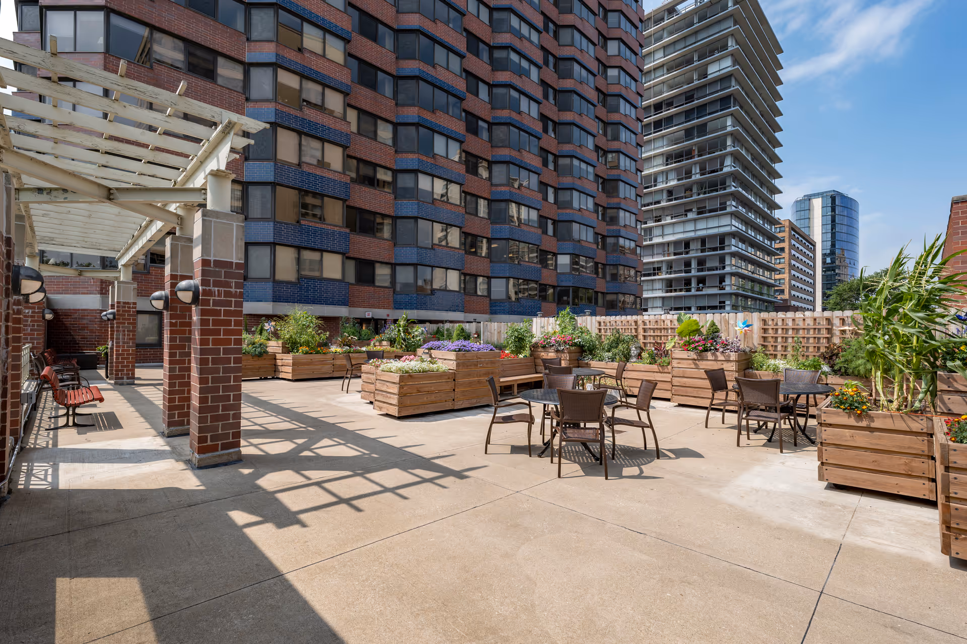 Rooftop outdoor patio with tables and chairs, wooden planter boxes, a pergola and surrounding high-rise buildings.