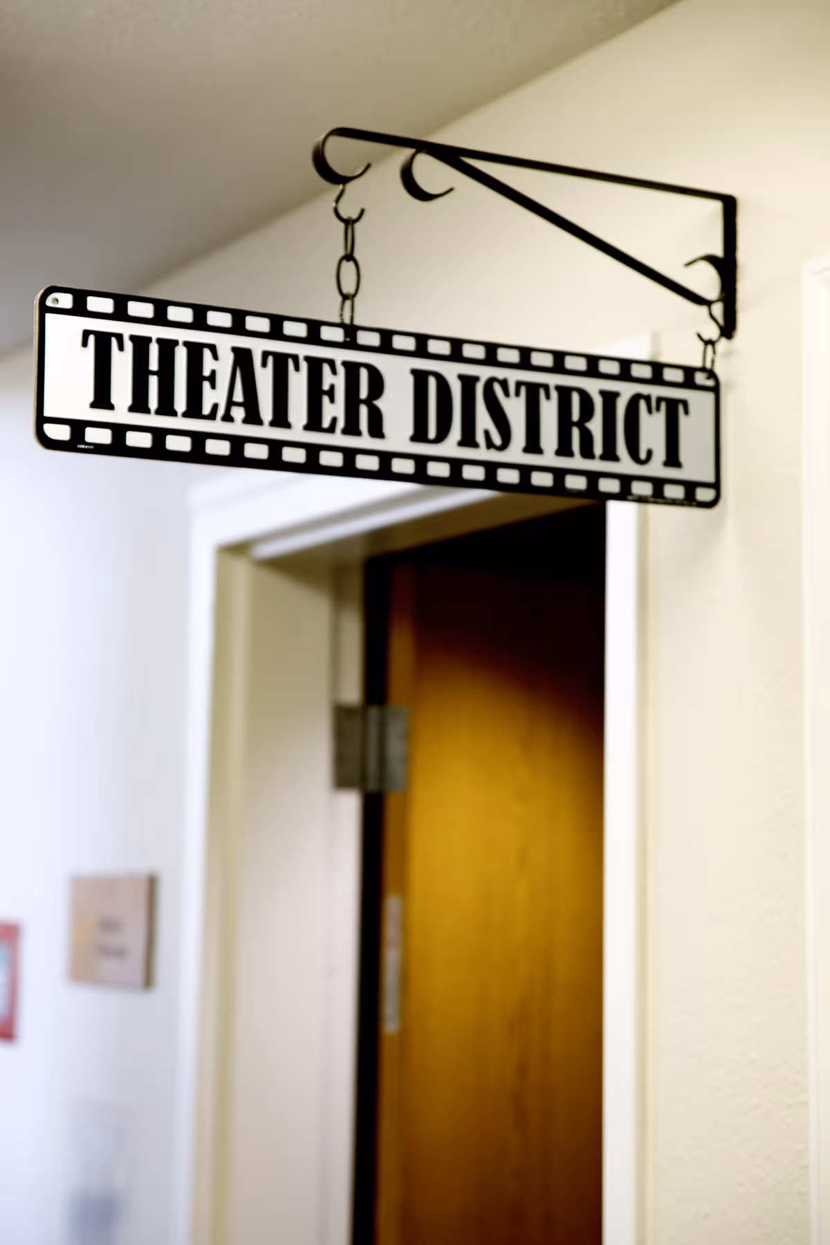 A hanging sign inside a building that reads 'THEATER DISTRICT' with a filmstrip border design, mounted on a decorative black metal bracket above a doorway.