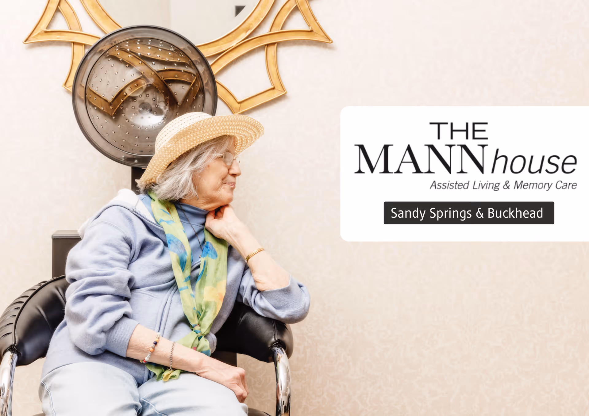 An elderly woman wearing a straw hat, glasses, a blue sweater, and a colorful scarf is sitting in a salon chair under a hair dryer in a room with beige wallpaper. She is looking to the side with her hand resting on her neck.