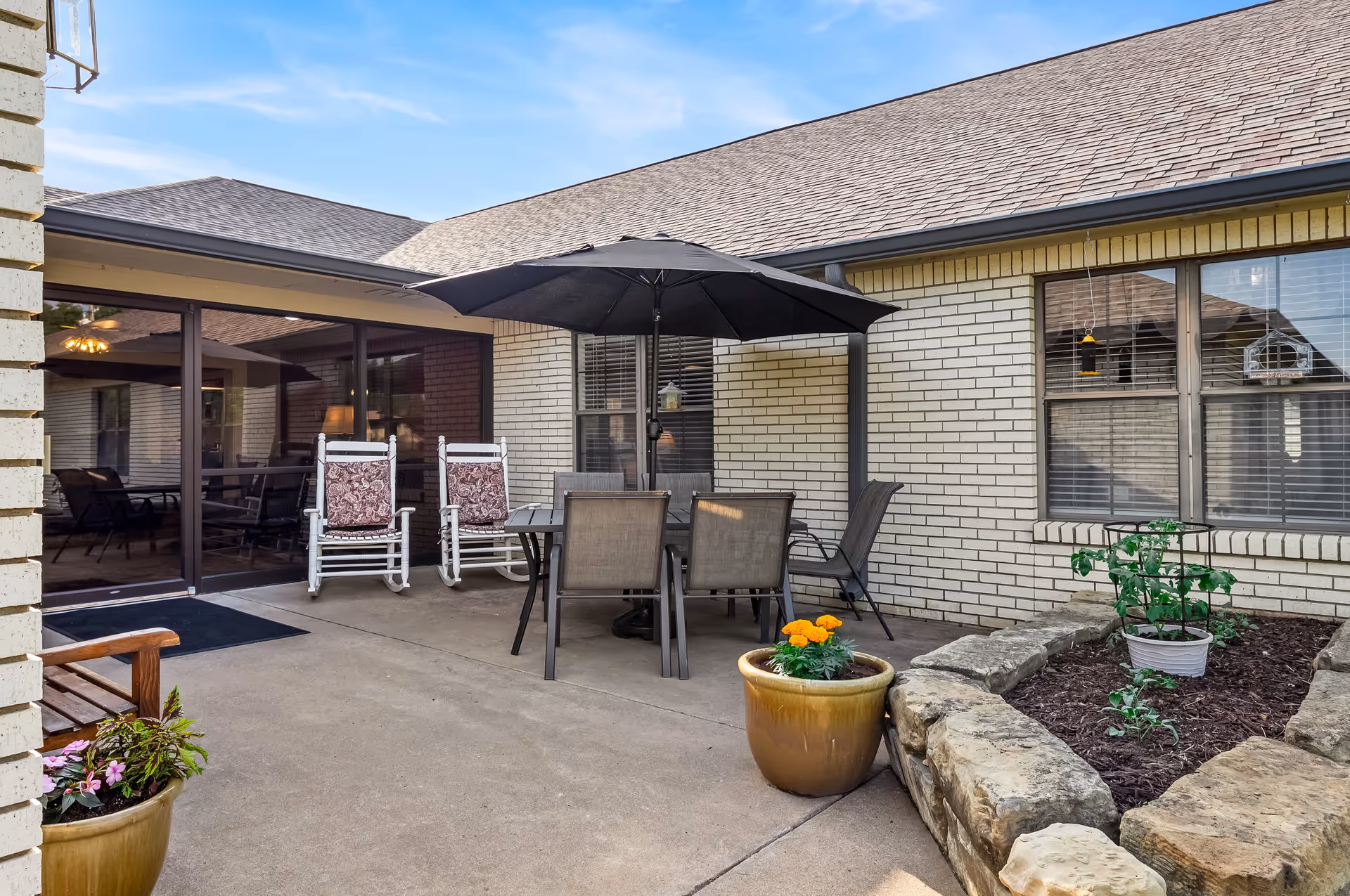 Outdoor patio area with a table and four chairs under a large black umbrella, two white rocking chairs with patterned cushions, potted plants, and a small garden bed with stone edging. The patio is adjacent to a building with light-colored brick walls and large windows.