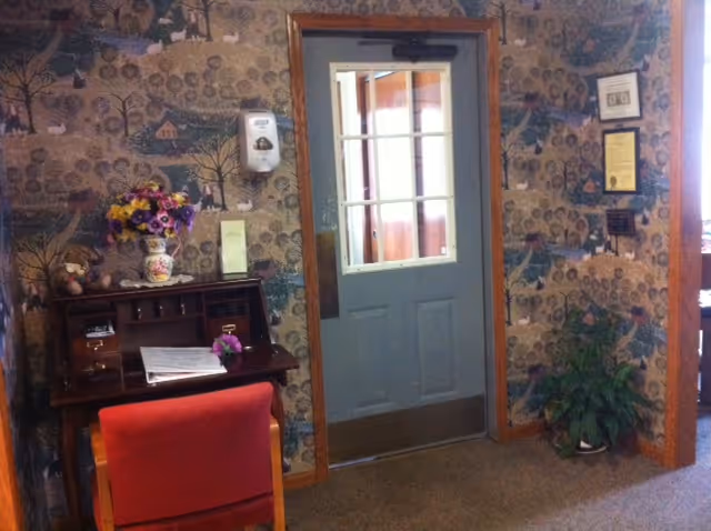 Interior view of a room with patterned wallpaper featuring trees and houses. A wooden desk with a vase of colorful flowers and some papers is positioned against the wall. A red chair is in front of the desk. A gray door with glass panes is centered in the image, and a green potted plant is on the floor to the right. A hand sanitizer dispenser is mounted on the wall to the left of the door.