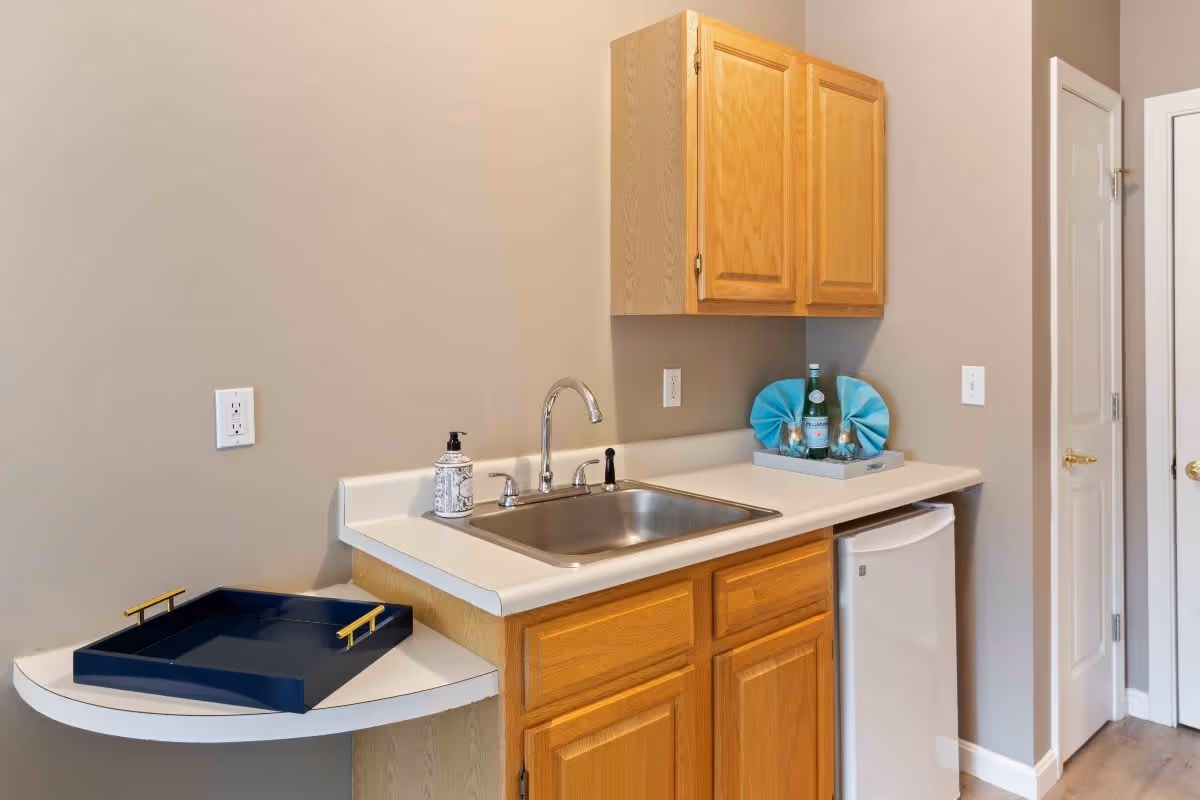 A small kitchenette area with wooden cabinets, a stainless steel sink, a white countertop, a mini refrigerator, a soap dispenser, and a decorative tray with a bottle and blue napkins. There is also a small white wall-mounted shelf with a dark blue tray on it.