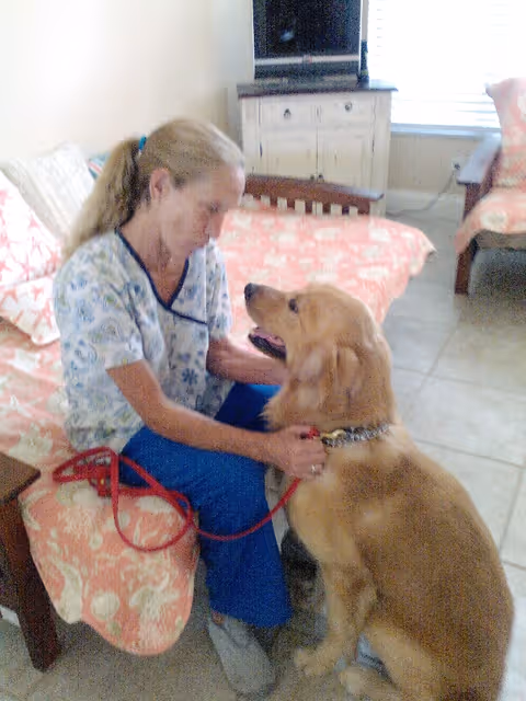 A woman in a floral scrub top and blue pants is sitting on a bed with a coral and white patterned bedspread, holding a red leash and interacting affectionately with a golden retriever dog sitting on the floor in a room with tiled flooring and light-colored walls.