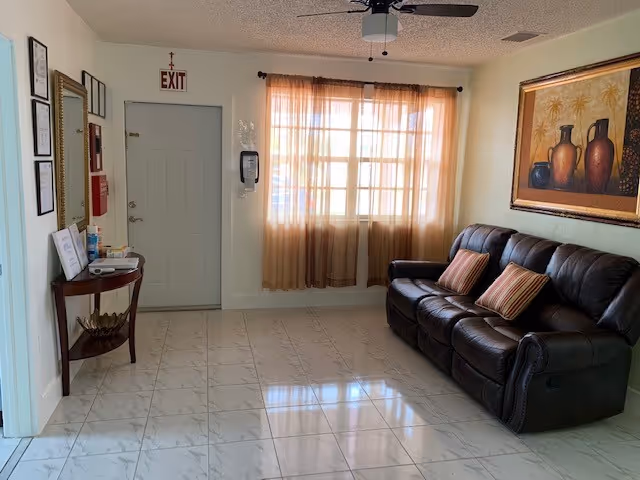 A small waiting or sitting area with a dark brown leather couch adorned with two striped pillows, a large framed painting of vases on the wall above the couch, a window with sheer beige curtains letting in natural light, a ceiling fan, a small wooden table with hand sanitizer and other items near the door, and an exit sign above the door.