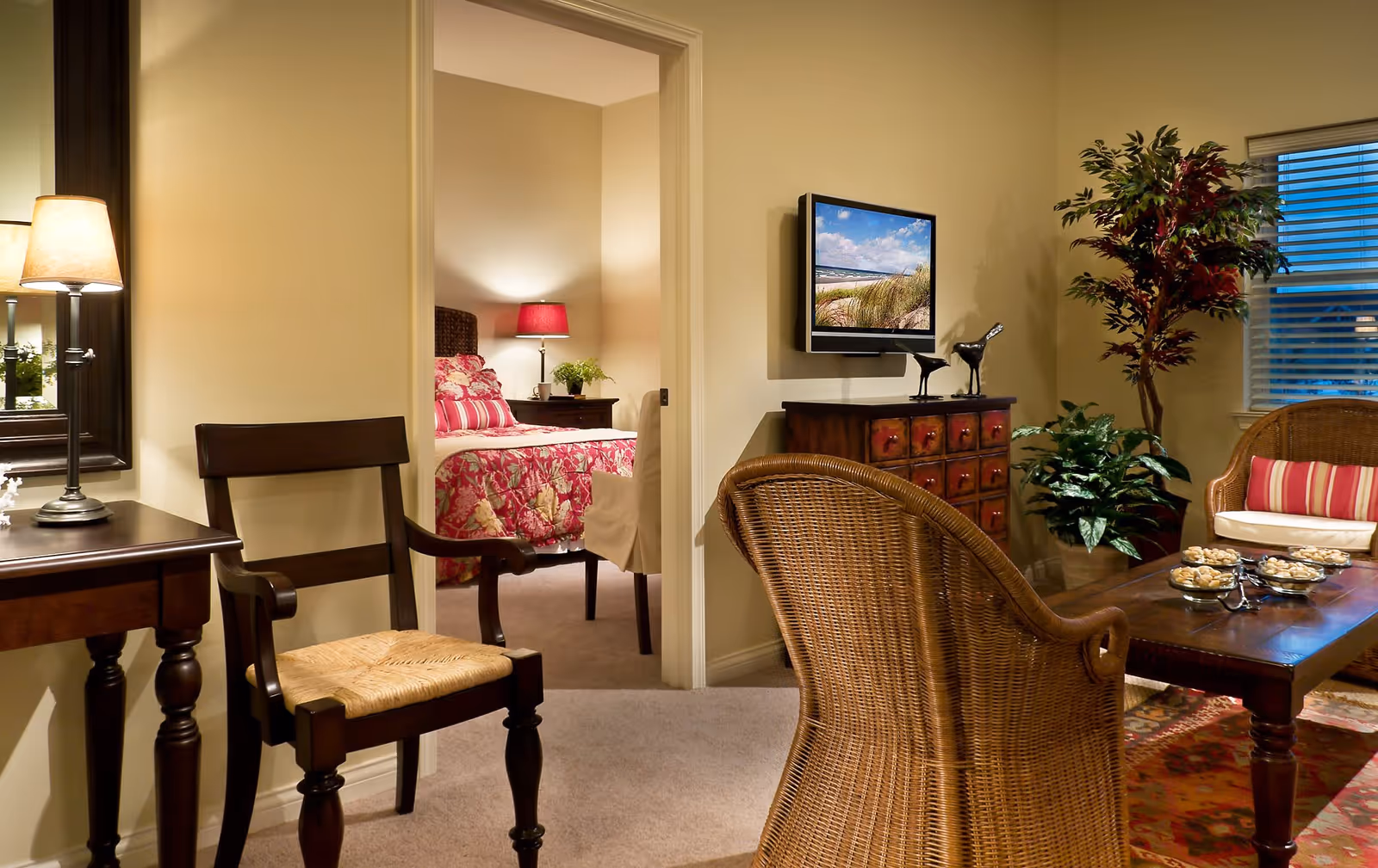 A cozy living room with wicker chairs, a wooden coffee table with decorative bowls, a chest of drawers with two bird figurines, a wall-mounted TV displaying a beach scene, and a large potted plant near a window with blinds. Through an open doorway, a bedroom is visible with a bed covered in a floral pink and white bedspread, a nightstand with a lamp and a small plant.
