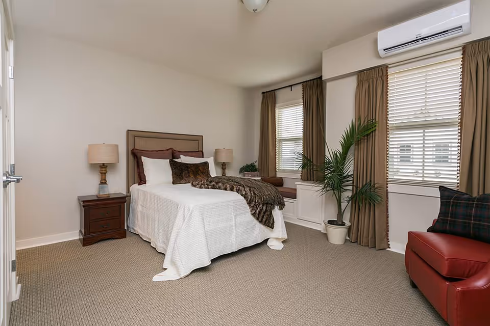 A neatly arranged bedroom with a bed covered in white bedding and multiple pillows, flanked by two wooden nightstands each with a lamp. The room has beige carpet, two windows with brown curtains, a potted plant, a red armchair with a plaid pillow, and a window seat beneath the windows. An air conditioning unit is mounted above one window.