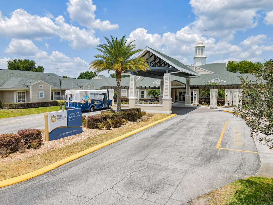 Exterior view of Solaris Healthcare East Orlando nursing and rehabilitation center showing the entrance driveway with a covered drop-off area, a palm tree, landscaped bushes, and a shuttle bus parked near the building under a partly cloudy sky.