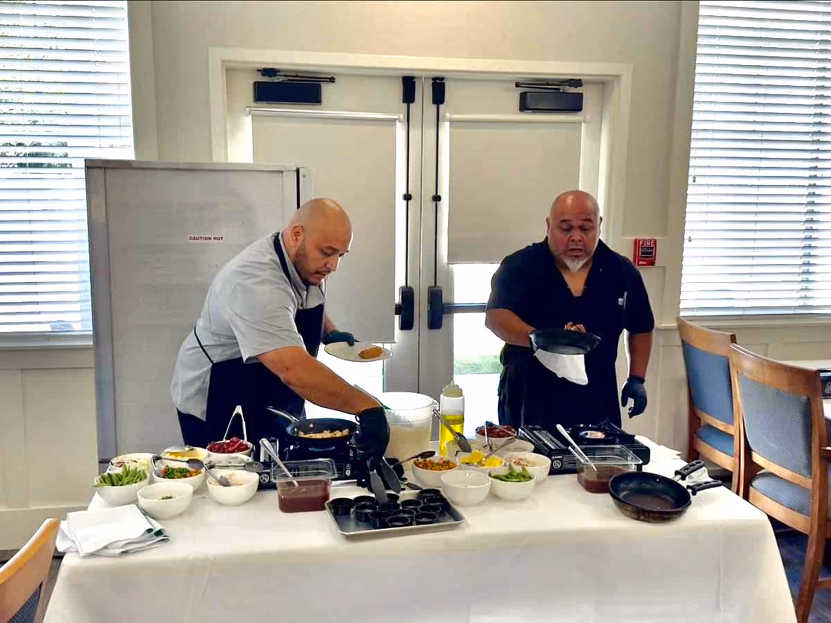 Two chefs preparing food at a table covered with a white tablecloth, with various bowls of ingredients and cooking utensils. They are in a bright room with large windows and closed blinds, and there are wooden chairs with blue cushions nearby.