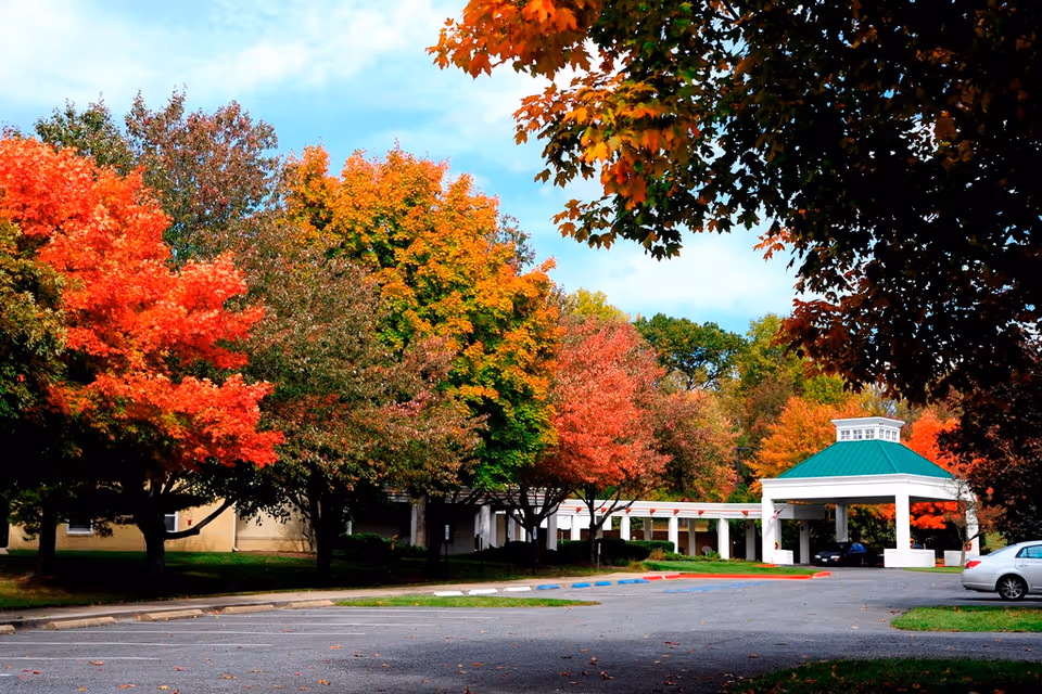 Entrance of a senior living community with a covered porte-cochère, parking area, and trees showing autumn foliage.