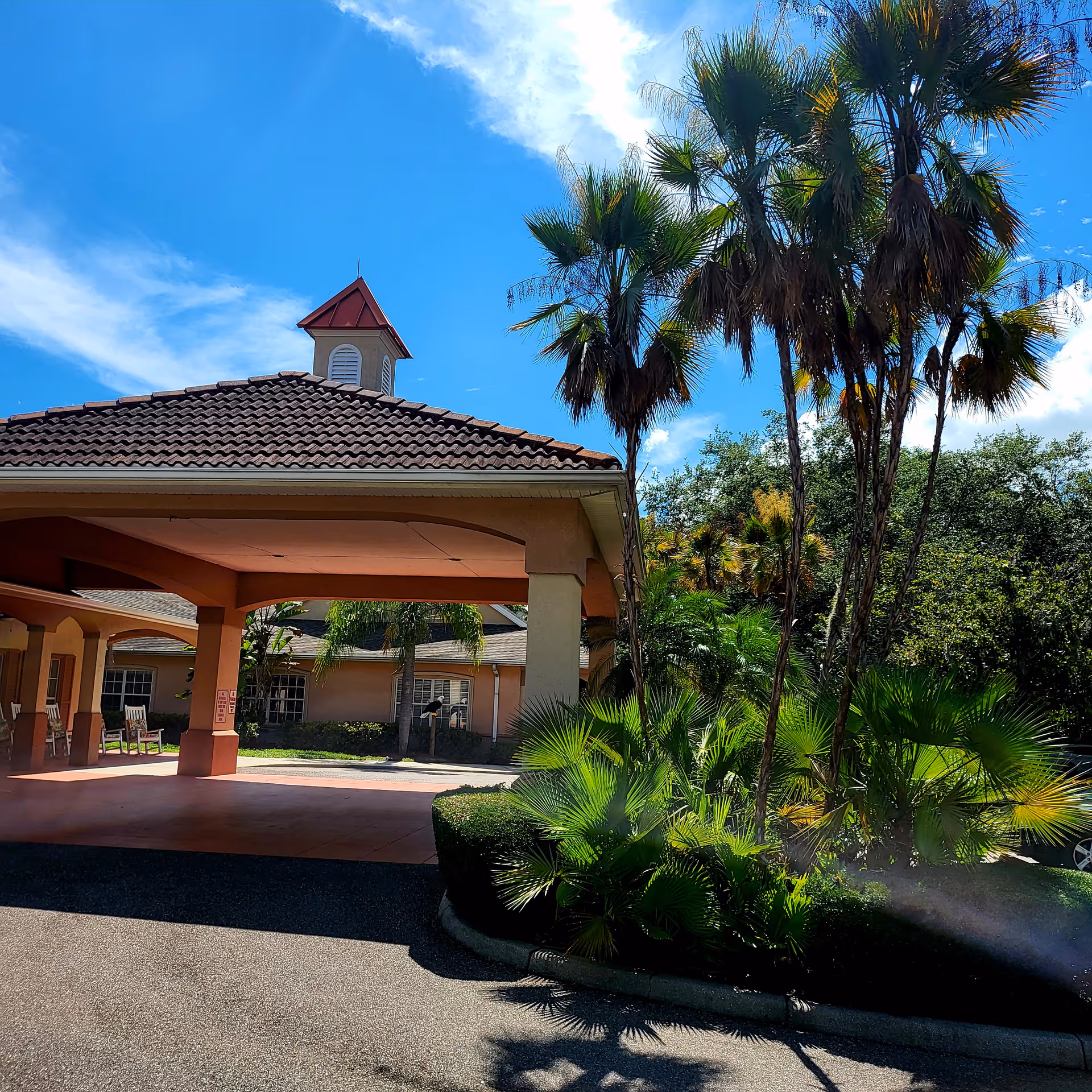 Exterior view of a building with a covered entrance featuring a tiled roof and a small tower with a red top. The building is surrounded by lush green palm trees and bushes under a bright blue sky with some clouds.