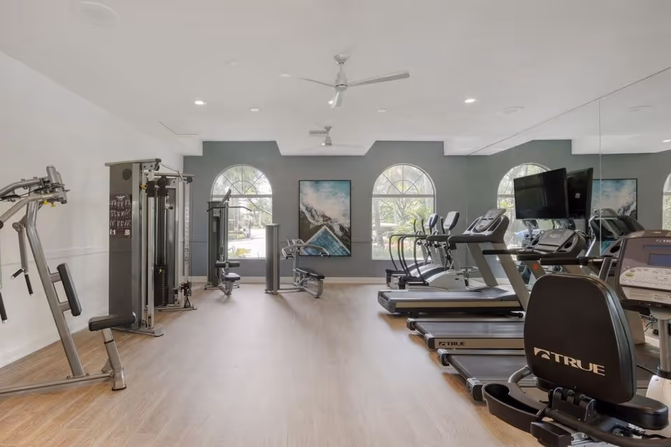 Bright fitness room with treadmills, weight machines, and large arched windows.
