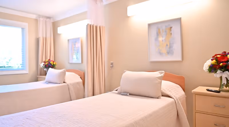 A bright and clean healthcare center bedroom with two single beds, each with white bedding and a large pillow. There are beige curtains between the beds, a window on the left side, and a nightstand with a colorful flower arrangement and a remote control. Soft wall lights and framed abstract artwork are visible above each bed.