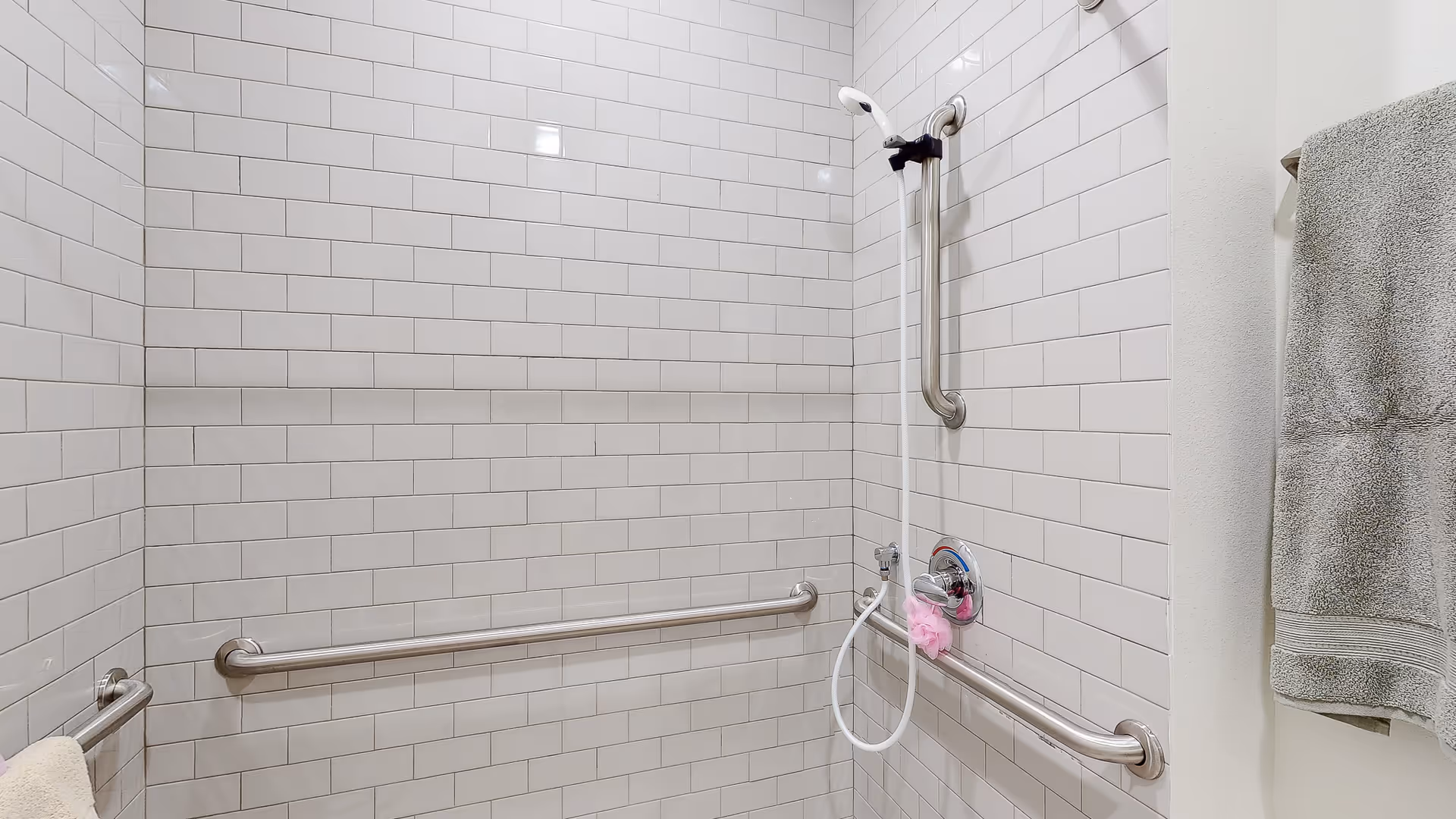 A tiled shower area with white subway tiles, equipped with stainless steel grab bars on three walls. There is a handheld showerhead attached to a vertical grab bar, and a pink bath sponge hanging from the shower control. A gray towel is hanging on a towel rack outside the shower area.