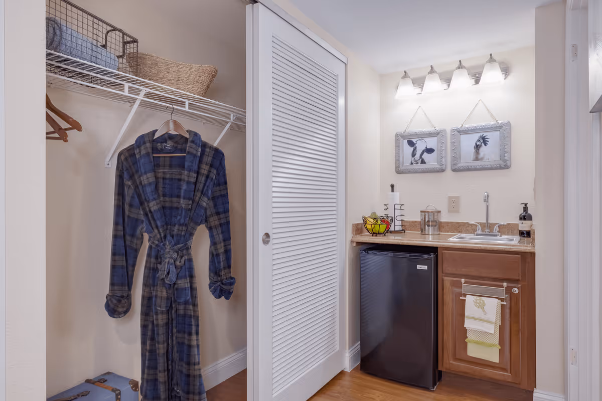 Small kitchenette with a sink, mini fridge and wooden cabinet beside an open closet holding a hanging blue plaid robe.