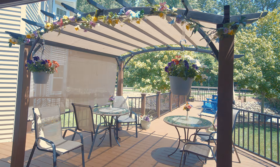 Outdoor patio area with a pergola adorned with hanging flower pots and floral decorations. The patio has two round glass-top tables, each with two chairs, and small flower vases on the tables. The area is surrounded by a black metal railing and overlooks a green lawn and trees.