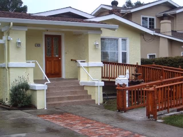 Front exterior view of a yellow senior living facility building with a wooden door, steps leading up to the entrance, white railings, a window with blinds, and a wooden ramp with railings on the right side.