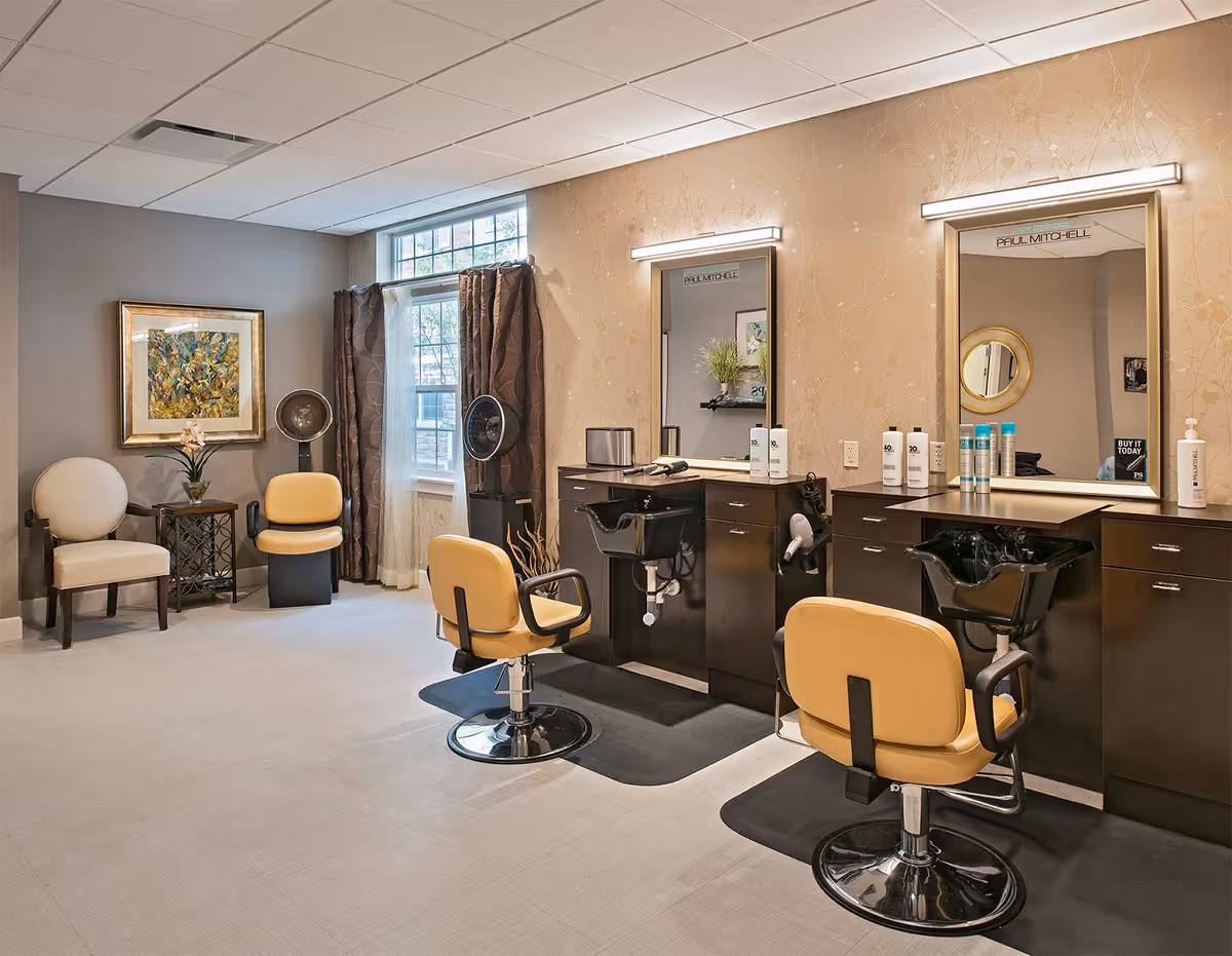 Interior of a salon area with two styling stations featuring large mirrors, salon chairs, and hair washing sinks. The room has beige walls with subtle floral patterns, a window with curtains, a framed painting, and additional seating with chairs and a small table.
