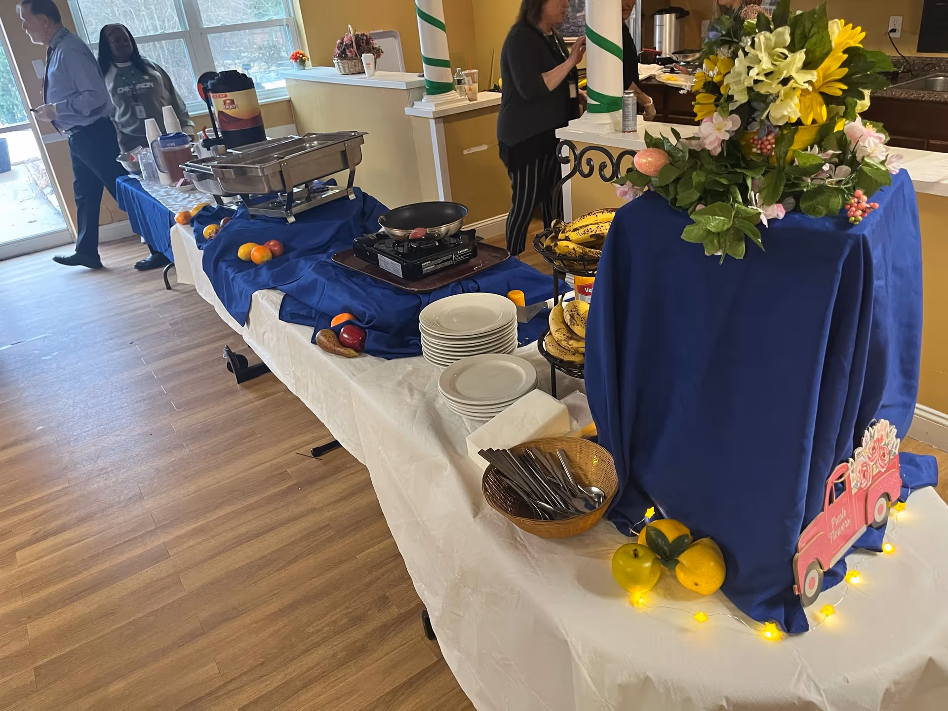A buffet table set up in a dining area with plates, serving trays, fruit, a floral centerpiece and staff standing in the background.