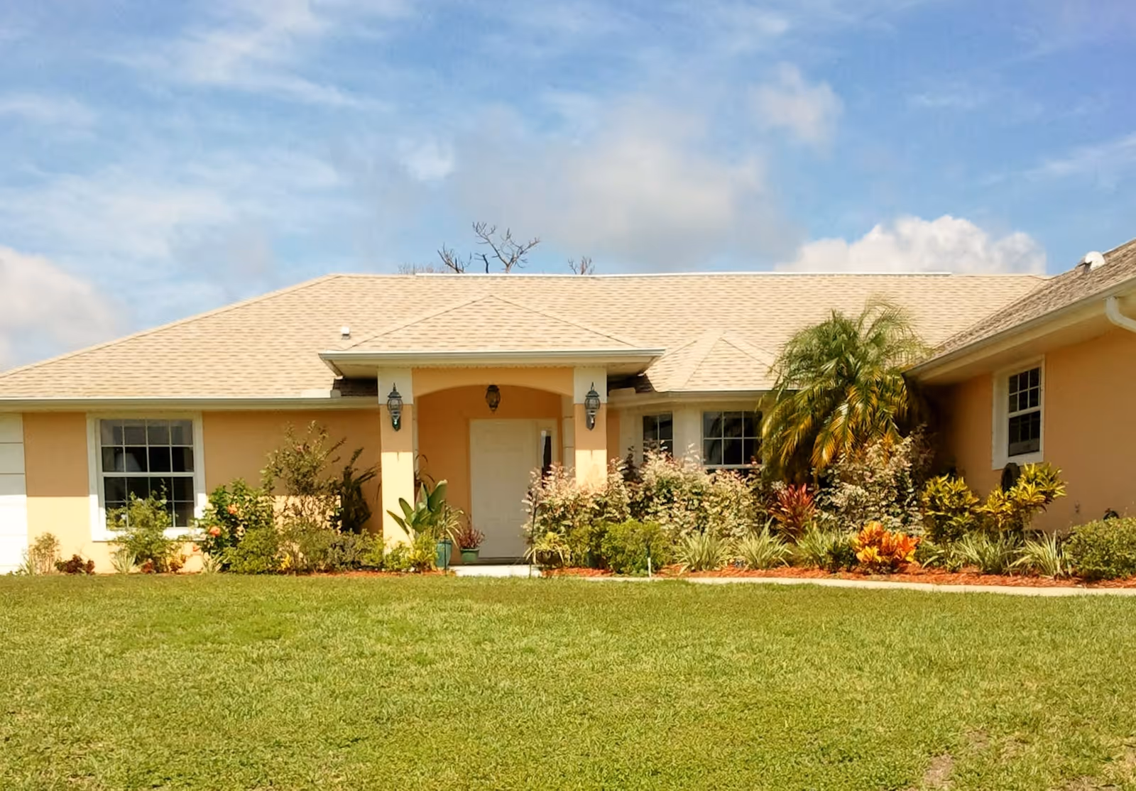 Single-story building with a beige exterior and a light-colored roof, surrounded by a well-maintained lawn and various shrubs and plants under a partly cloudy sky.