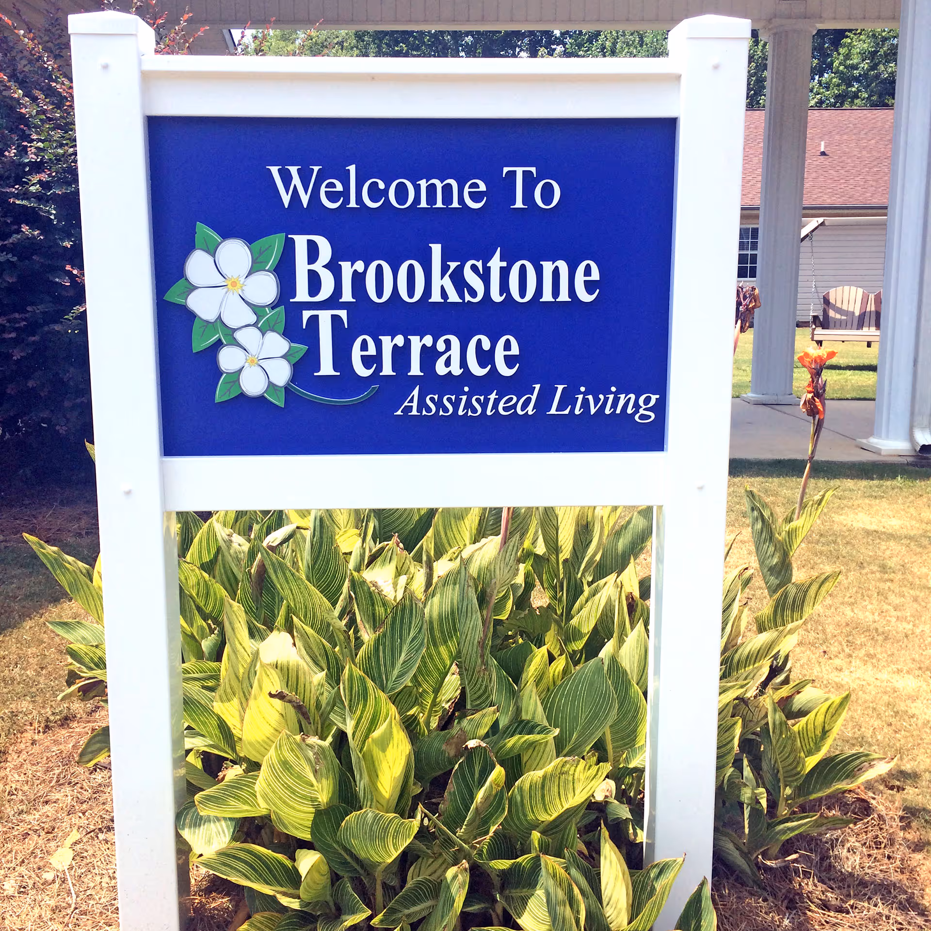A white-framed blue sign with white flowers that reads 'Welcome To Brookstone Terrace Assisted Living' standing in front of green leafy plants with a building and lawn visible in the background.