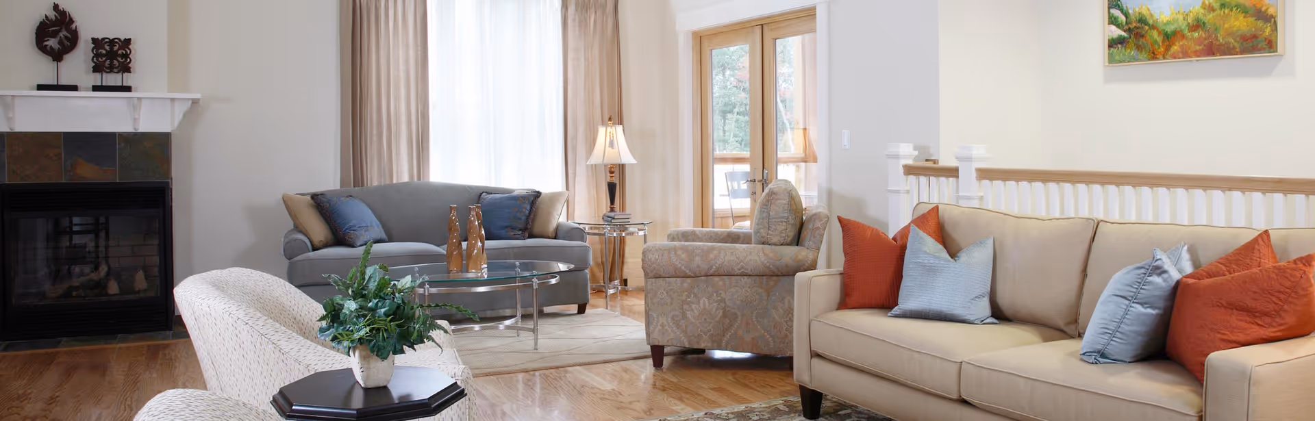 A cozy living room with a beige sofa adorned with red and blue pillows, a patterned armchair, a gray loveseat with blue and beige pillows, a glass coffee table with decorative items, a side table with a lamp, a fireplace with a white mantle, and large windows with beige curtains letting in natural light.
