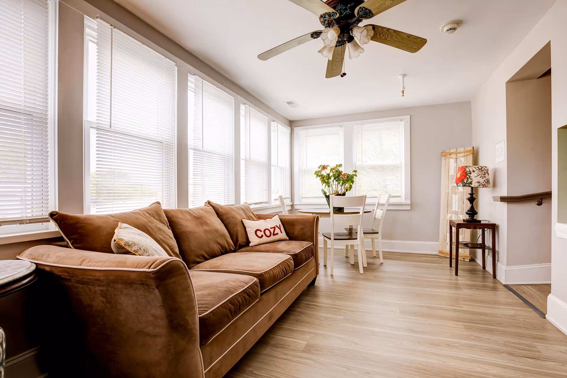 A bright and cozy living room area with a brown sofa featuring a pillow with the word 'COZY'. The room has large windows with white blinds allowing natural light to fill the space. There is a small dining table with four white chairs and a plant centerpiece near the windows. A ceiling fan with lights is mounted on the ceiling, and a small side table with a decorative lamp is positioned near an open doorway.